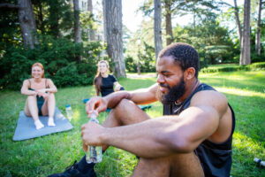 A man with a beard is sitting on a mat in a park is drinking water with two other people