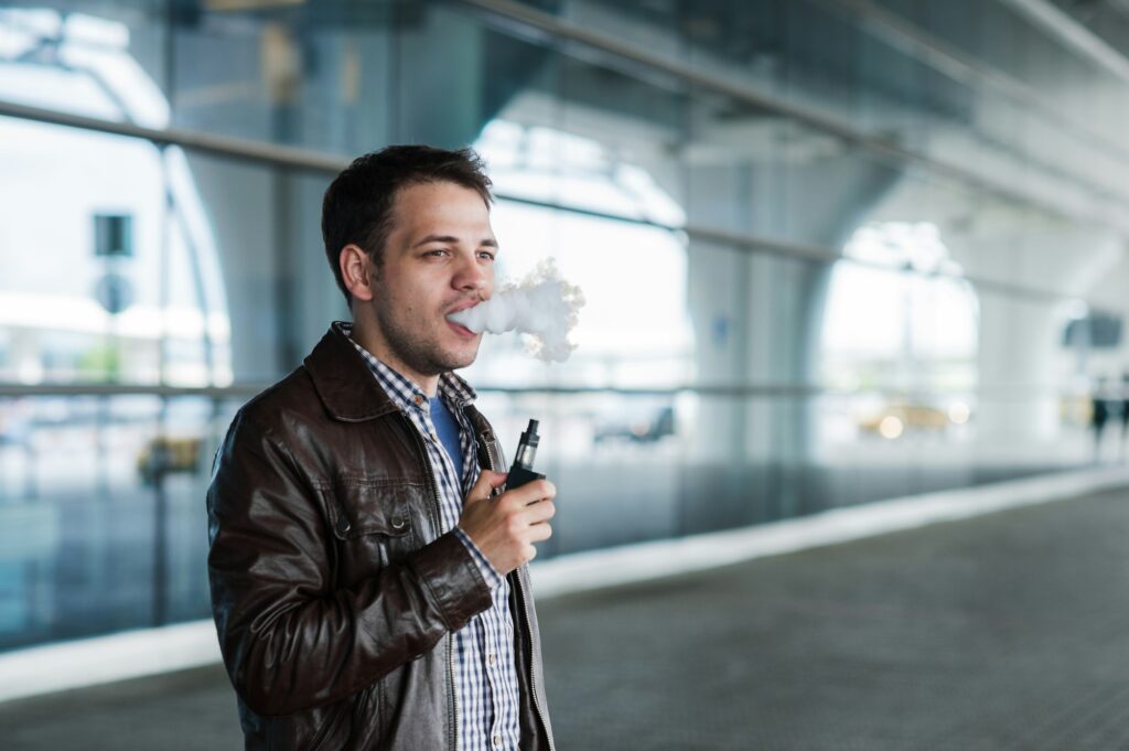 Man with a bristle smoking e-cigarette vaporizer box mode outdoors near the airport terminal before